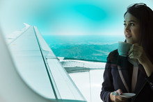 Young Business Woman Looking Forward, With A Cup Of Coffee In Hand. Selective Focused On The Airplane Wing And Having Blurry Mountian In Background. Air Traveling And Transportation Concept. 