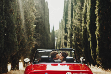 look from behind at wedding couple kissing in a red cabrio which stands between tall trees somewhere