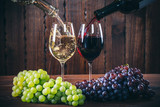 Pouring red and white wine into the  wine glasses with a bunch of red and white grapes against wooden background