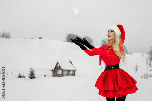Fotografie, Obraz Beautiful sexy blonde woman wearing a red dress and a Santa's hat on a background of mountains and snow