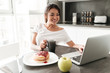 © Drobot Dean - Cheerful young woman sitting at the kitchen using laptop