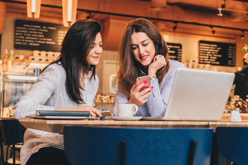 Two young happy women are sitting in cafe at table in front of laptop, using smartphone and laughing. On table paper notebook and cup of coffee. Girls are blogging, working, studying, learning online.