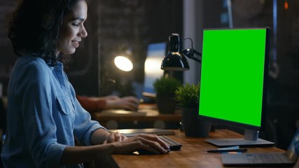 Poster - Beautiful Female Office Employee at Her Desk Works on a Mock-up Green Screen Personal Computer. Her Colleague Sits Beside. Creative Office Evening. RED EPIC-W 8K Camera.