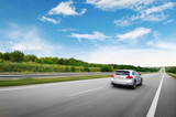 Fototapeta Sport - A silver car driving fast on the countryside road against sky with clouds