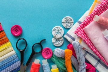 Multicolored threads, scissors, buttons, fabric and various sewing accessories on a blue background with copy space flat lay