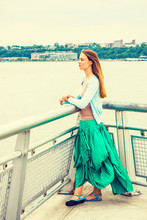 I Waiting For You. American College Student Wearing Light Blue Cardigan, Green Skirt, Sandals, Standing By Metal Fence By Hudson River In New York, Opposite New Jersey, Holding Book, Looking Forward..