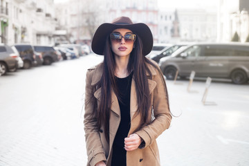 Wall Mural - Outdoor portrait of young beautiful fashionable woman with long hair walking in the street. Model looking aside, wearing stylish classic beige coat, hat, accessories. Female fashion, beauty concept