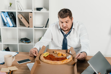 Wall Mural - overweight businessman looking at pizza while sitting in office