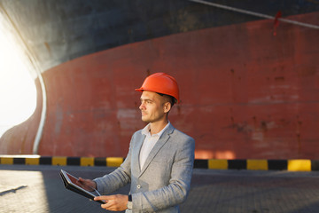 Handsome unshaven successful business man in gray suit, protective construction orange helmet holding tablet, standing in sea port against cargo rusty ship background. Male with gadget in sunny day