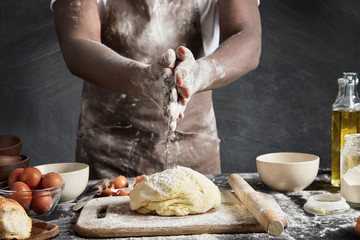 Cropped image of unrecognizable male chef wears dirty apron, prepares dough for making loaf, uses different ingredients, isolated over black chalk background. Talented African American cook.