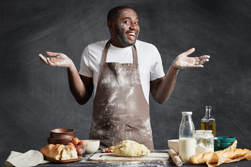 Uncertain male chef shrugs shoulders, has happy expression, doesn`t know what to bake, prepares dough, stands near kitchen table, isolated over black chalk background. Hesitant dark skinned baker