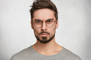 Wall Mural - Headshot of strict annoyed aggressive man with beard, mustache, looks seriously through round spectacles, frowns face in dissatisfaction, expresses negative emotions, isolated over white concrete wall