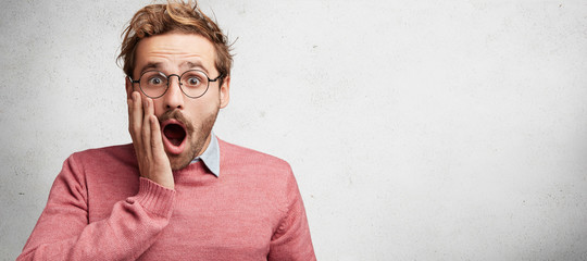 Wall Mural - Horizontal shot of amazed stupefied bearded male wonk wears spectacles, looks with shocking expression, keeps mouth widely opened, being frightened see car accident on street, isolated on white wall