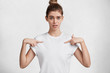 © WHstudio Leushin N - Puzzled beautful woman indicates at new white t shirt, bought oversized clothing, isolated over white studio background. Blue eyed female points at blank space on shirt for your advertisment or design