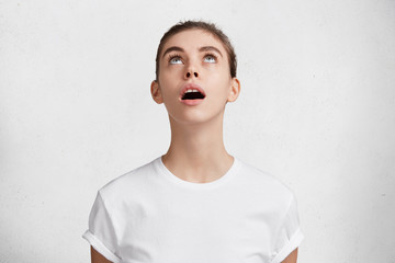 Wall Mural - Curious beautiful woman looks with intriguing expression upwards, notices something interesting, keeps mouth opened, dressed in white casual t shirt, isolated over concrete wall. Curiosity concept