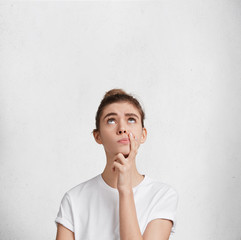 Wall Mural - Vertical shot of thoughtful female model being deep in thoughts, tries to decide what do on weekends, looks pensively upwards, isolated over white concrete wall with copy space for promotional text