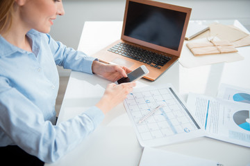 Poster - Top view smiling career girl writing message in phone while sitting at desk in modern apartment. Appliance and documents situating on it. Business concept