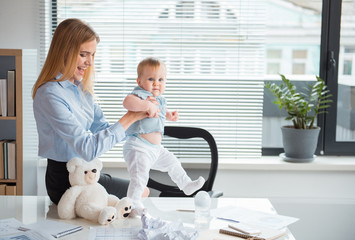 Side view smiling woman holding glad kid. Girl learning to go on desk in office. Occupation, family and balance concept