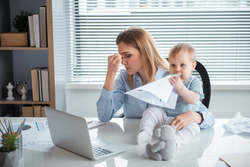 Poster - Weary mother working at desk while keeping baby on it. portrait of interested kid tasting paper. Job and family concept
