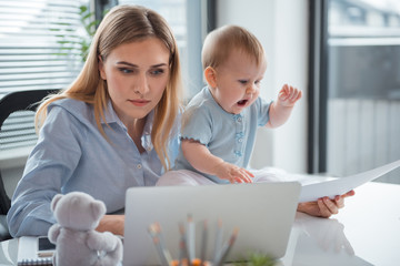 Portrait of serene young woman working at notebook computer while keeping document in hand. Yawning baby sitting near her and watching on it. Occupation and child concept