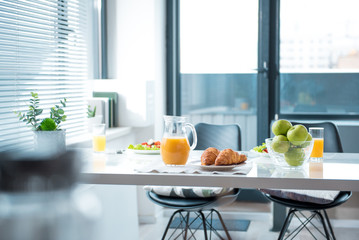 Fresh orange juice in jug, croissants and apples on table in kitchen. Plates with salad on background