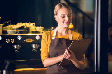 Wall Mural - Portrait of friendly young female barista reading information while standing in modern cafe. Occupation concept