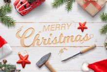Creative Chrtistmas Greeting With Carved Text On White Wooden Surface With Hammer And Chisel. Christmas Decorations Beside.