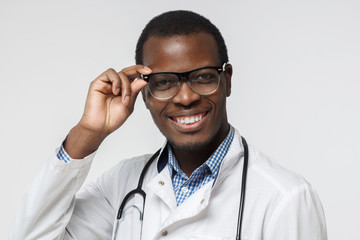 Wall Mural - Close up portrait of smiling african doctor holding eyeglasses, isolated on gray background