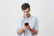 © Cookie Studio - Handsome joyful student with dark hair wearing blue shirt messaging, typing message, using free onlipe app on his smartphone, looking at screen with smile, posing against gray background.