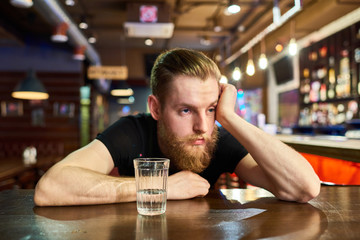 Front view portrait of drunk bearded man in pub sitting at table with glass of vodka looking away with sad expression