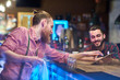 © Seventyfour - Side view portrait of modern bearded bartender talking to client at bar counter in pub, helping him choose drinks via digital tablet
