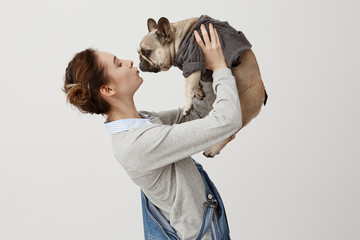 Horizontal shot of adult girl in jeans overalls kissing cute puppy while raising it in air. Young girl being in love with her dog clothed in trendy jumper. Display of affection