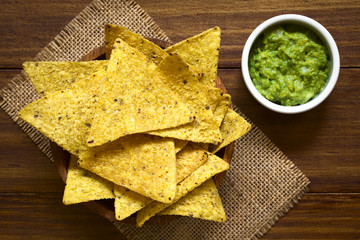 Corn tortilla chips in bowl with avocado dip on the side, photographed overhead on wood with natural light (Selective Focus, Focus on the top of the tortilla chips)