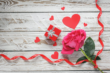 Wall Mural - Rose with red paper hearts and gift box and ribbon on white wooden background. Love symbol concept.