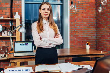 Confident female business owner standing at her work desk folding arms looking at camera in creative design studio