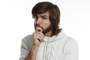 Portrait of unshaven young male hipster in stylish sweater with hood having suspicious or skeptical look, touching his chin and gazing at camera with his squinted eyes expressing doubt and disbelief