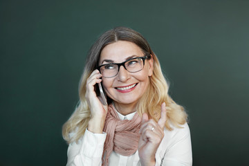 Wall Mural - Isolated portrait of cunning positive 60 year old blonde female in eyewear gesturing with index finger, smiling broadly and looking up while talking on cell phone. People, age and electronic devices