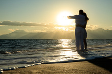 Young Sexy Couple In Love Is Walking At A Beautiful Sunset Beach At The Sea. Caribbean Style And A Mountain Range In Backround. Holding Hands And Pointing To The Landscape