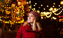 Outdoor Photo Of Young Beautiful Happy Smiling Girl Posing In Street. Festive Christmas Fair On Background. Model Wearing Stylish Winter Coat, Knitted Beanie Hat, Scarf.