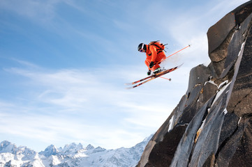 A professional skier makes a jump-drop from a high cliff against a blue sky leaving a trail of snow powder in the mountains