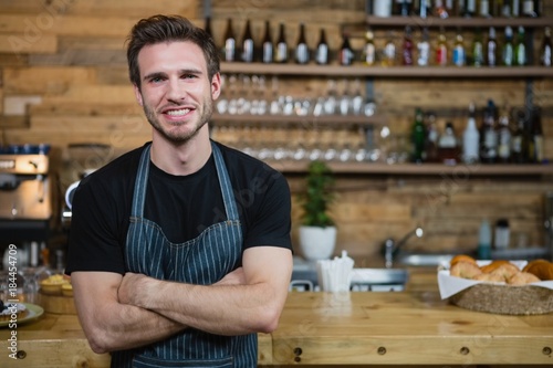 Cuadros en Lienzo Portrait of smiling waiter standing with arms crossed at counter