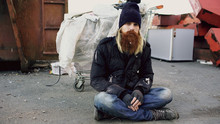 Portrait Of Young Bearded Homeless Man Sitting On A Sidewalk Near Shopping Cart Ang Garbage Container During Cold Winter Day