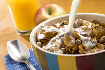 Milk pouring over a bowl of cereal with a spoon, apple and orange juice in the background.