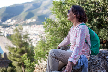 Wall Mural - beautiful elderly traveler woman sits on a stone and admires the sea from a height. Travel and leisure in retirement, active seniors