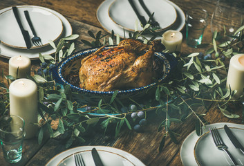 Whole roasted chicken with garlic for Christmas eve celebration decorated with olive tree branch over rustic wooden table background with plates and candles. Holiday table decoration set concept