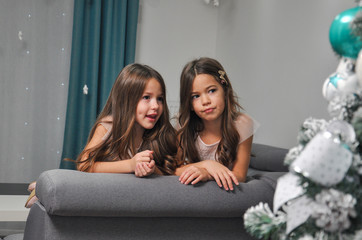 Two girls on sofa next to a Christmas tree talking and play on mobile phones. Sisters in living room waiting a New Year