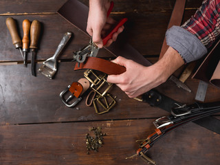 Leather master working holding a hole punch and a piece of leather. On brown wooden table scattered with tools and accessories.