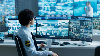 In the Security Control Room Officer Monitors Multiple Screens for Suspicious Activities, He Drinks from a Mug. He's Surrounded by Monitors and Guards Port of National Importance.