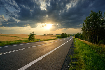Wall Mural - empty asphalt road leading around golden cornfields in rural landscape at sunset with dramatic cloud