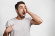 © wayhome.studio  - Portrait of scared amazed young man with stubble, keeps mouth widely opened, looks with terrified expression, indicates at blank copy space of t shirt, isolated over white concrete background